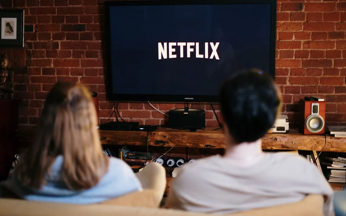Casal visto de costas assistindo a um serviço de streaming na televisão, em uma sala com parede de tijolos.