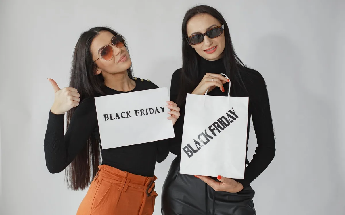 Duas mulheres de óculos escuros segurando placas e sacolas com os dizeres Black Friday.