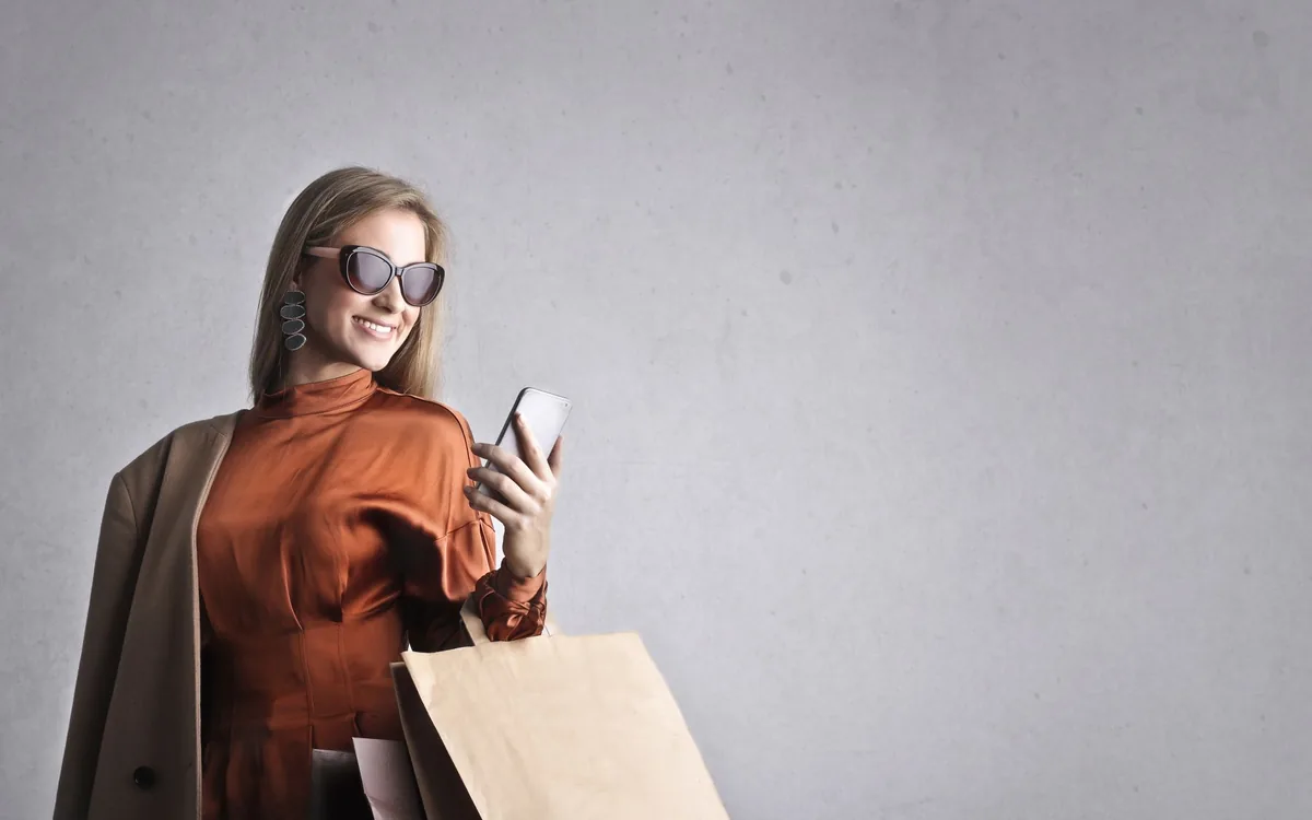 Mulher de óculos escuros olhando o celular enquanto segura sacolas de compras.