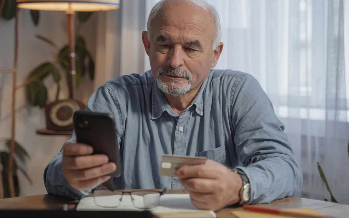 Homem mais velho sentado à mesa em casa, com o celular e um cartão de crédito nas mãos.