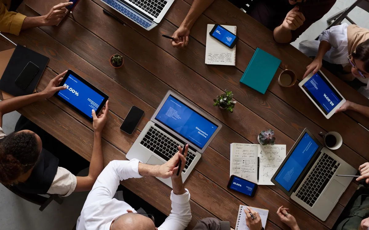 Vista de cima de uma mesa de reunião com notebooks, tablets e pessoas trabalhando juntas.
