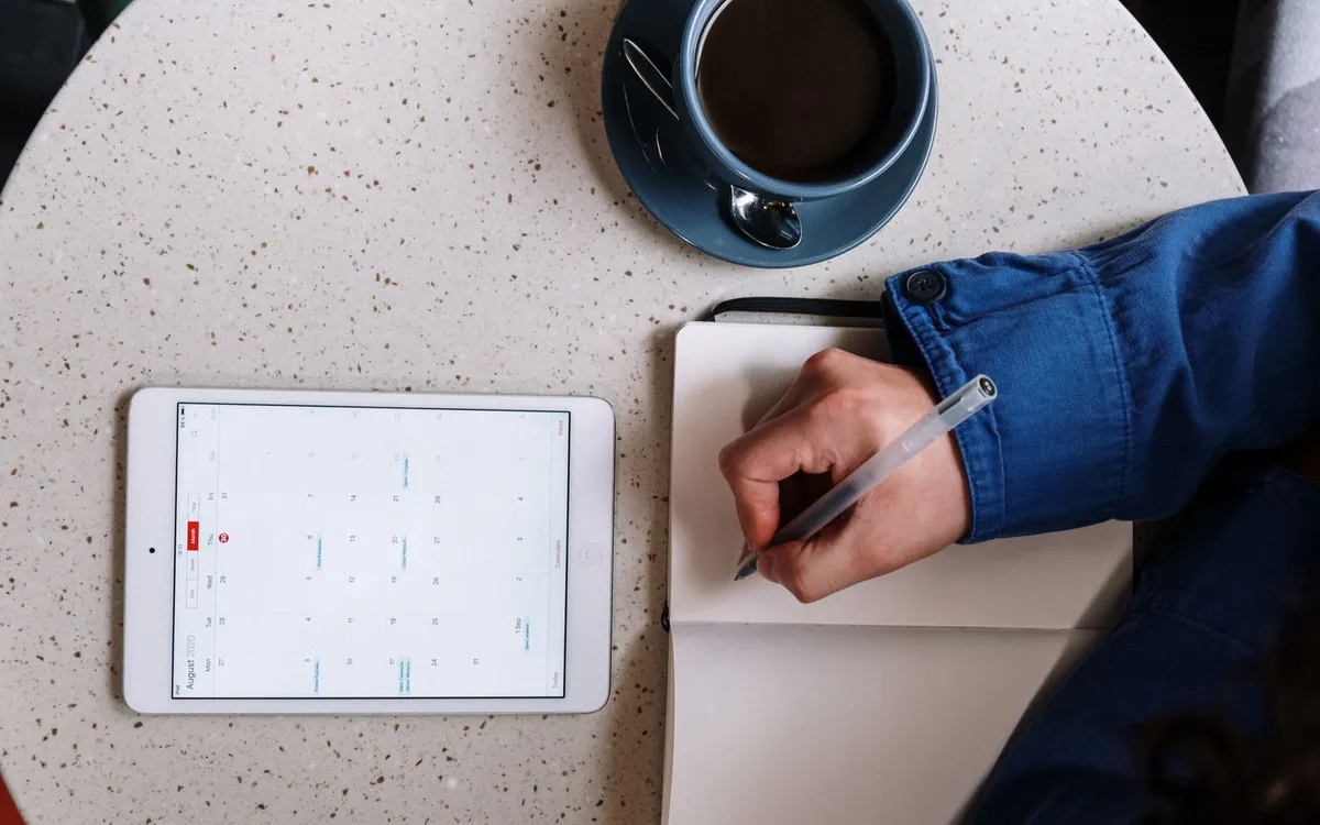 Mão escrevendo em um caderno ao lado de um tablet e uma xícara de café.