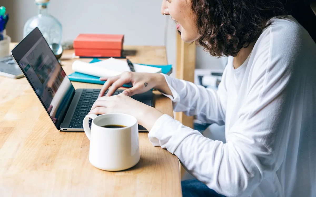 Mulher digitando em um notebook sobre uma mesa de madeira, ao lado de uma xícara de café.
