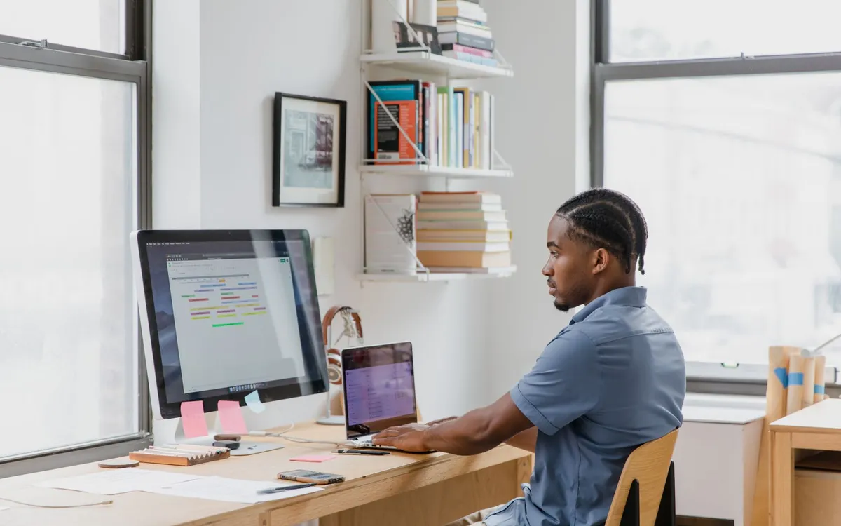 Homem trabalhando em um escritório doméstico com monitor, notebook e estante de livros.