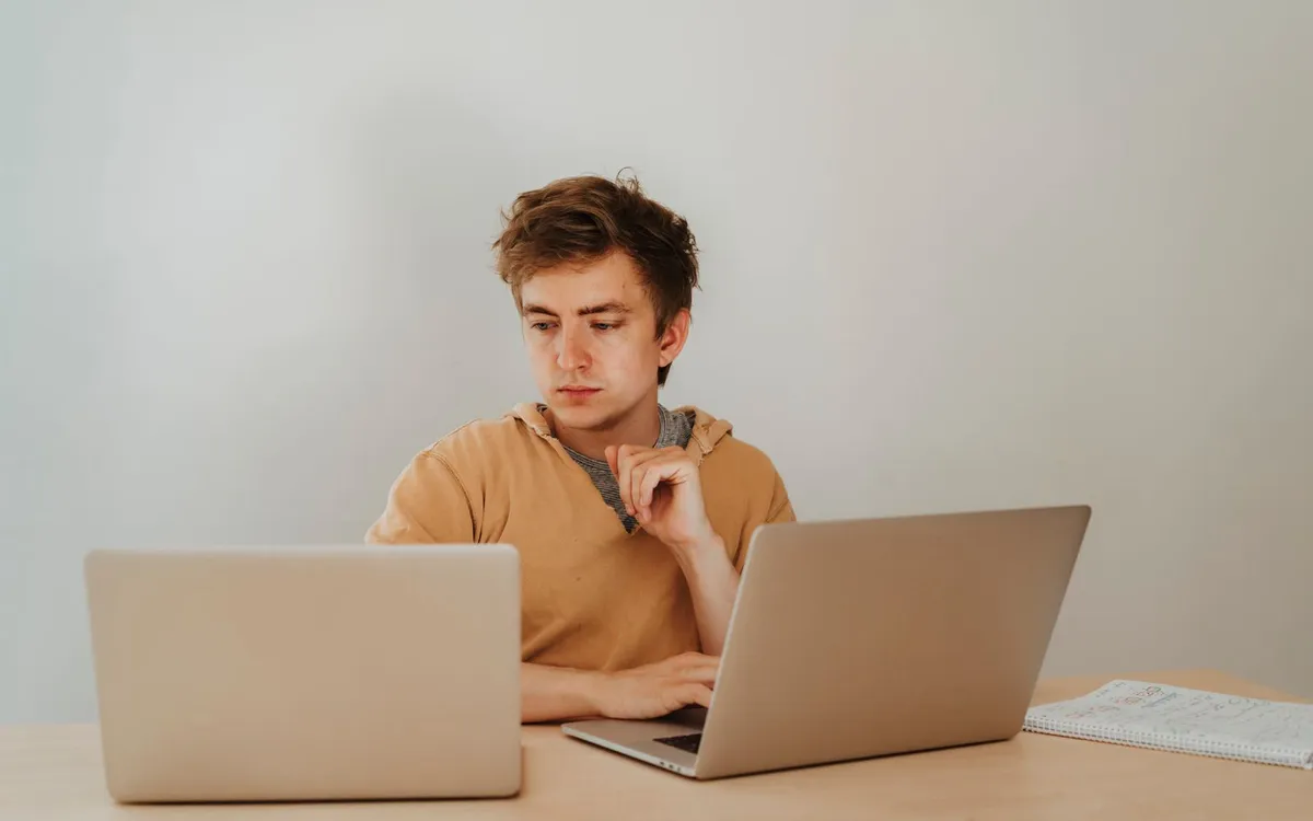 Jovem concentrado trabalhando em dois notebooks sobre uma mesa de madeira.