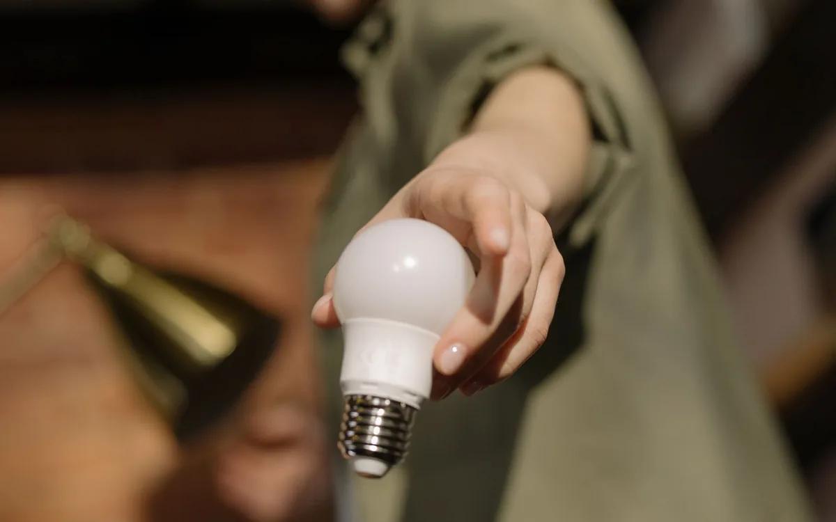 Mão segurando uma lâmpada LED branca em um ambiente de luz quente e fundo escuro.
