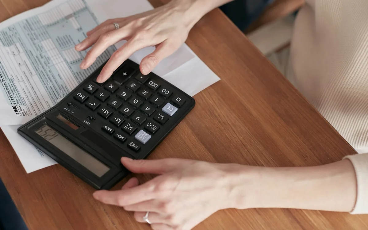 Close de mãos digitando em uma calculadora sobre contas e papéis, em uma mesa de madeira.