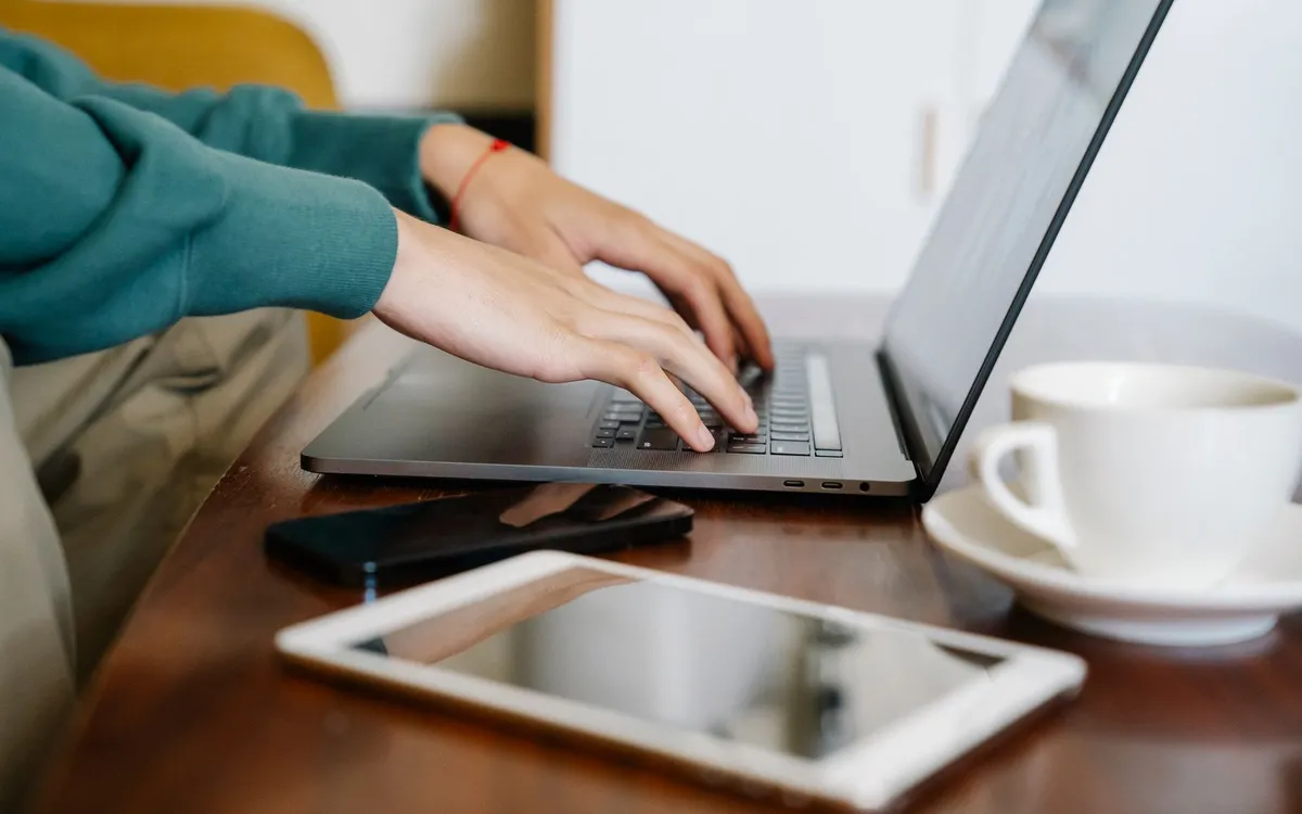 Mãos digitando em um notebook, com um tablet e uma xícara de café ao lado.