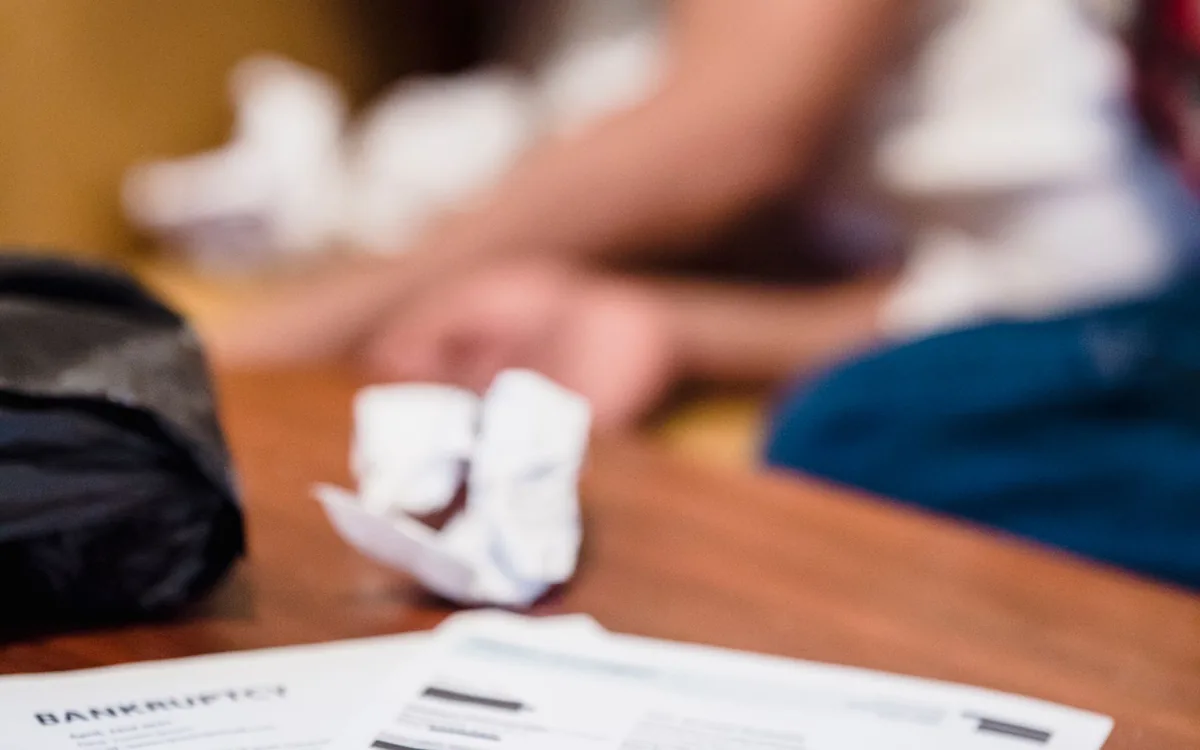 Contas vencidas sobre uma mesa, com um homem de cabeça baixa ao fundo.