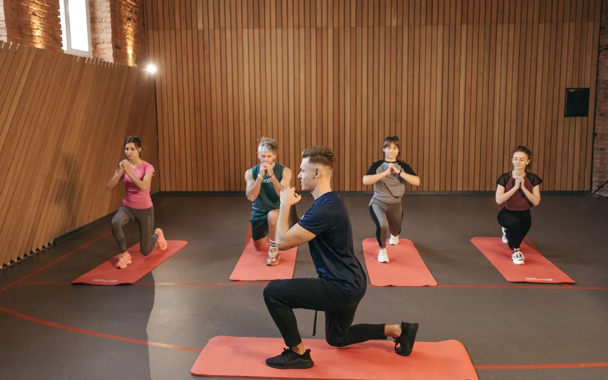 Aula coletiva em estúdio com alunos em tapetes laranja, fazendo avanços guiados pelo instrutor.