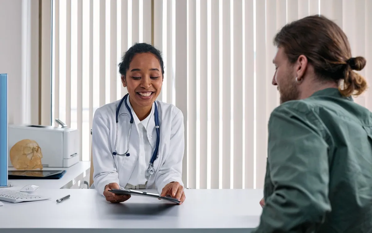 Médica sorrindo à mesa de consultório enquanto conversa com um paciente.