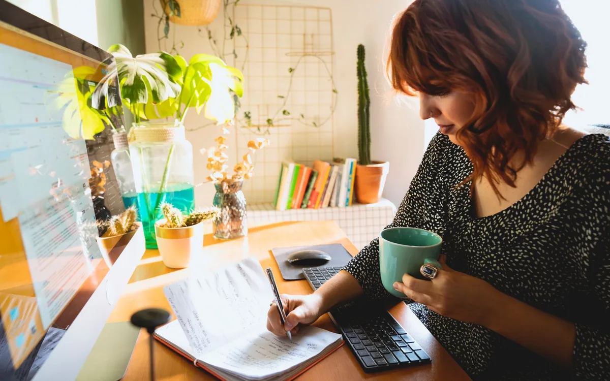 Mulher de cabelo ruivo escrevendo em um caderno em uma mesa com planta e caneca.