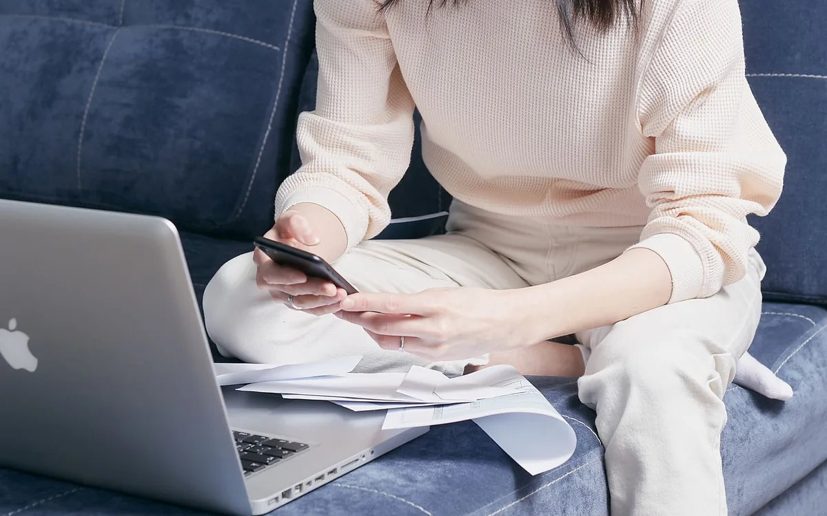 Mulher no sofá usando o celular, com notebook e papéis ao lado, organizando contas em casa.