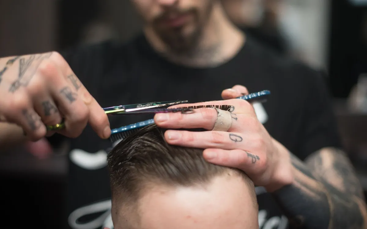 Barbeiro de braços tatuados cortando o cabelo de um cliente com tesoura e pente.
