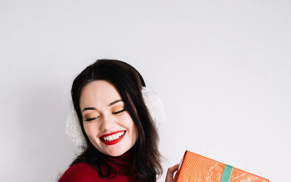 Mulher de suéter vermelho sorrindo de olhos fechados e segurando uma caixa de presente embrulhada.