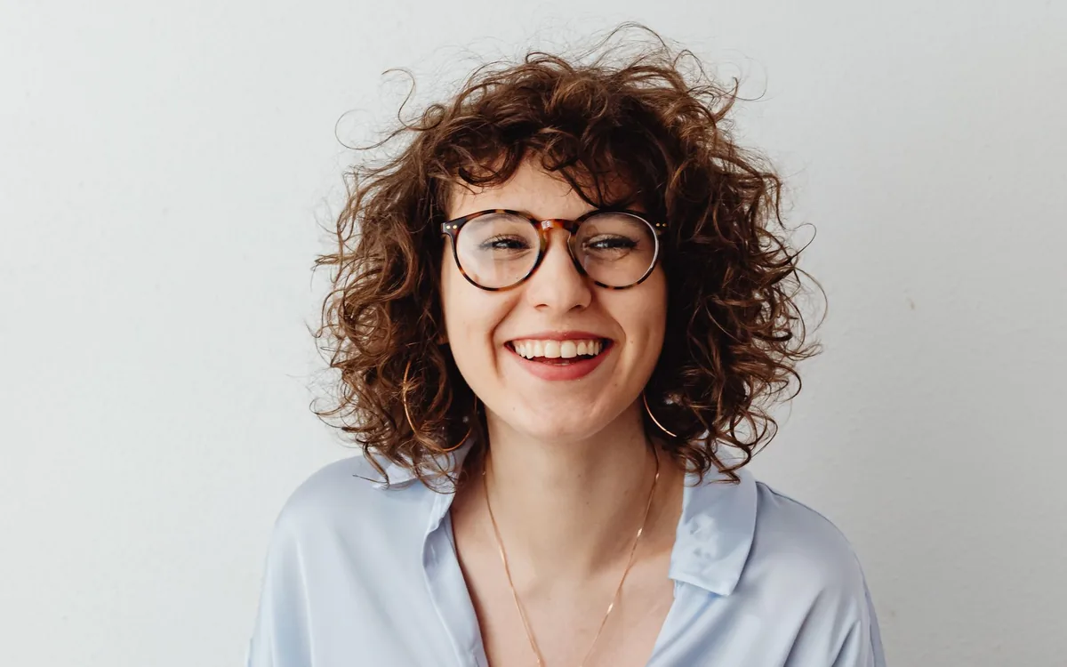 Mulher de cabelo cacheado e óculos sorrindo, sentada com dinheiro nas mãos.