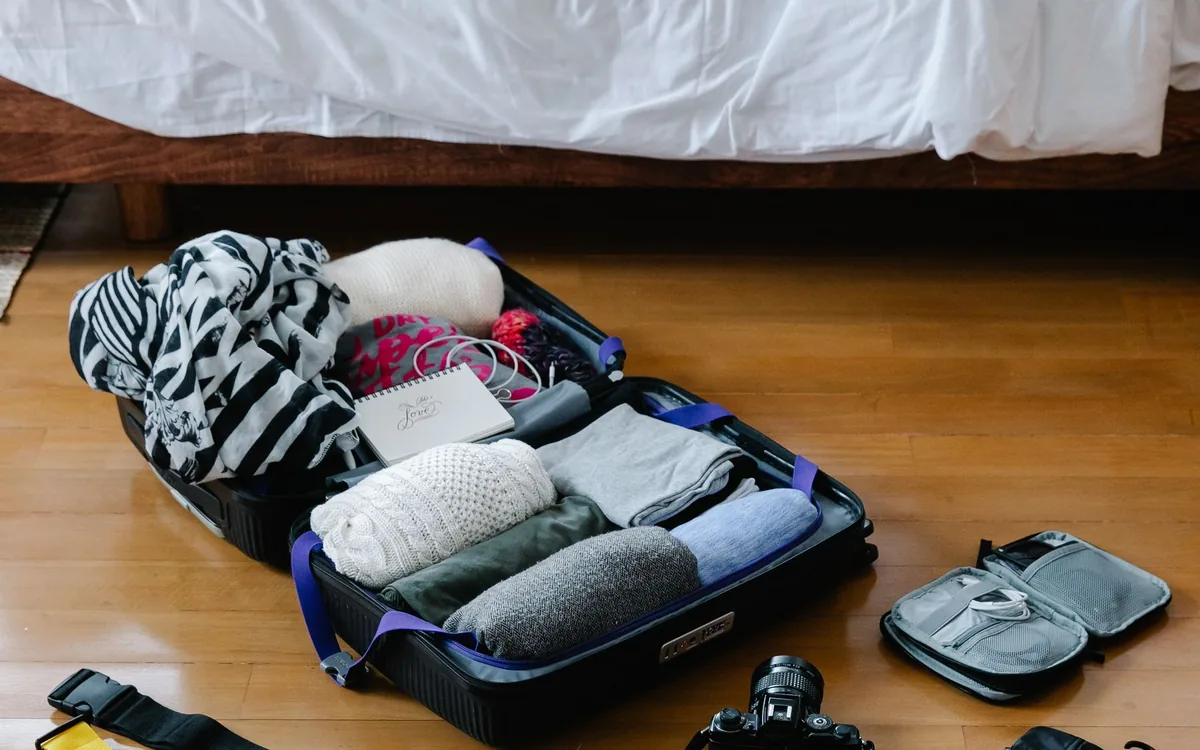 Mala aberta com roupas dobradas e itens de viagem sobre um piso de madeira.