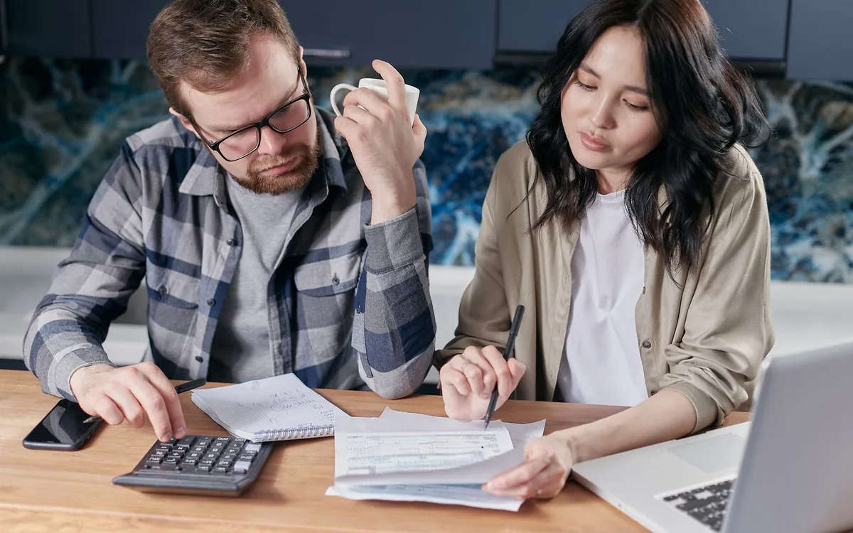 Casal com calculadora e papéis sobre uma mesa de madeira, anotando as contas da casa.