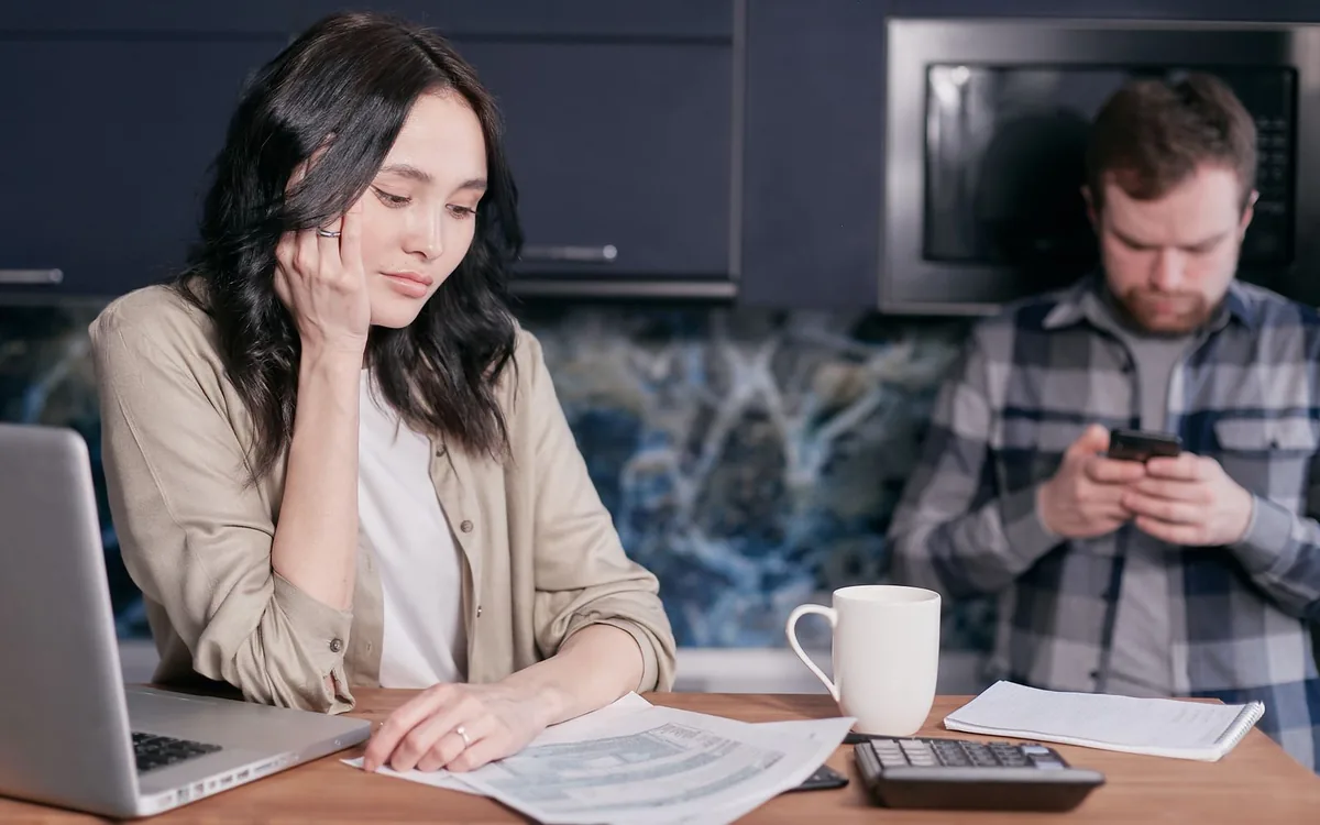 Mulher com a cabeça apoiada na mão ao lado de um homem que usa o celular, à mesa da cozinha.