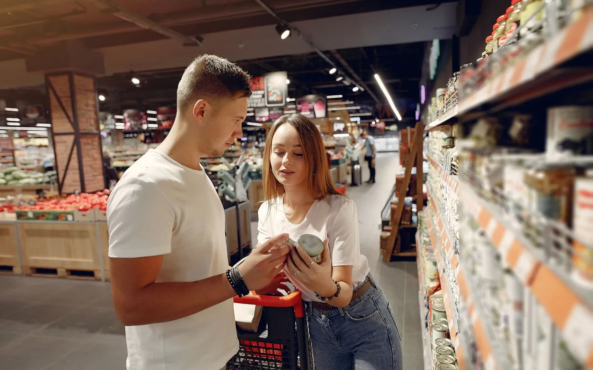 Casal em um corredor de supermercado olhando para um item, empurrando o carrinho.