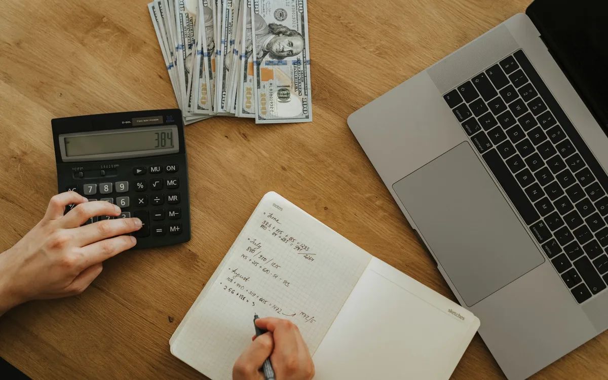 Mãos usando uma calculadora ao lado de um caderno, notas de dinheiro e um notebook sobre uma mesa de madeira.