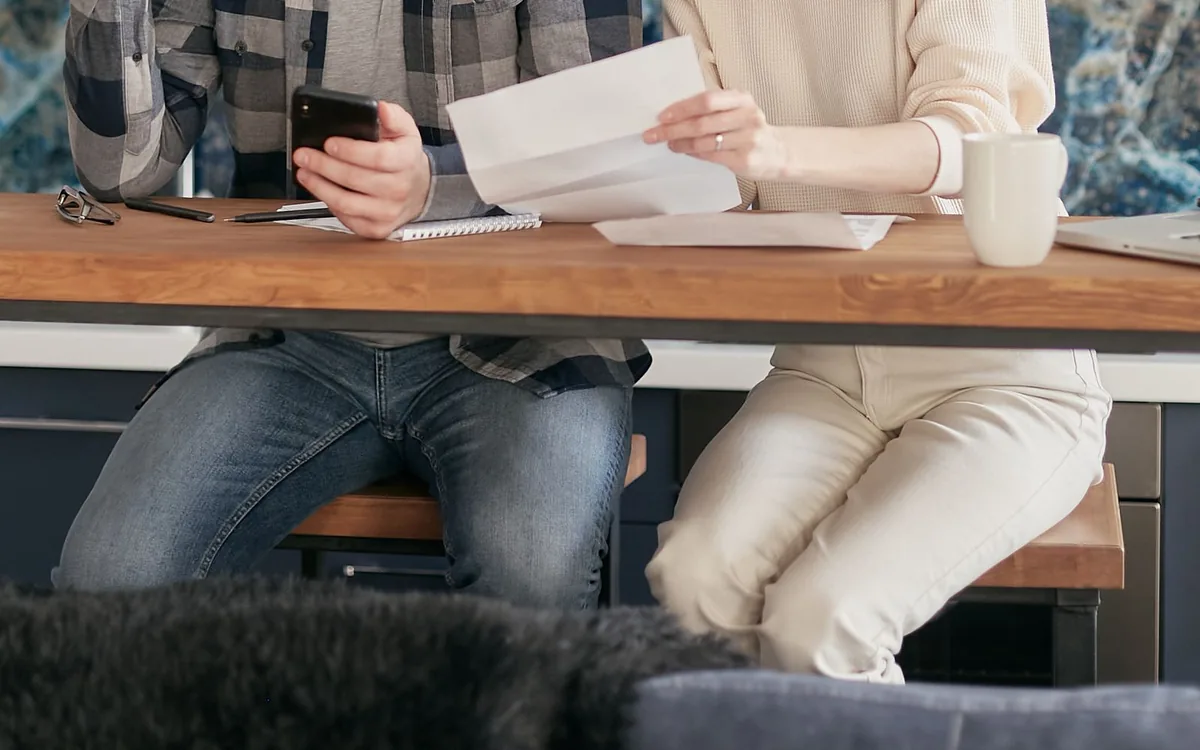 Casal à mesa da cozinha conferindo um papel e o celular, de frente para a bancada.