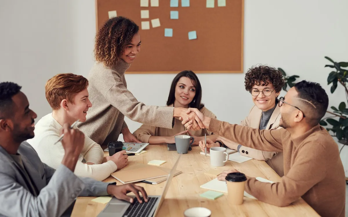 Equipe diversa apertando as mãos em torno de uma mesa de reunião, com post-its em um mural ao fundo.
