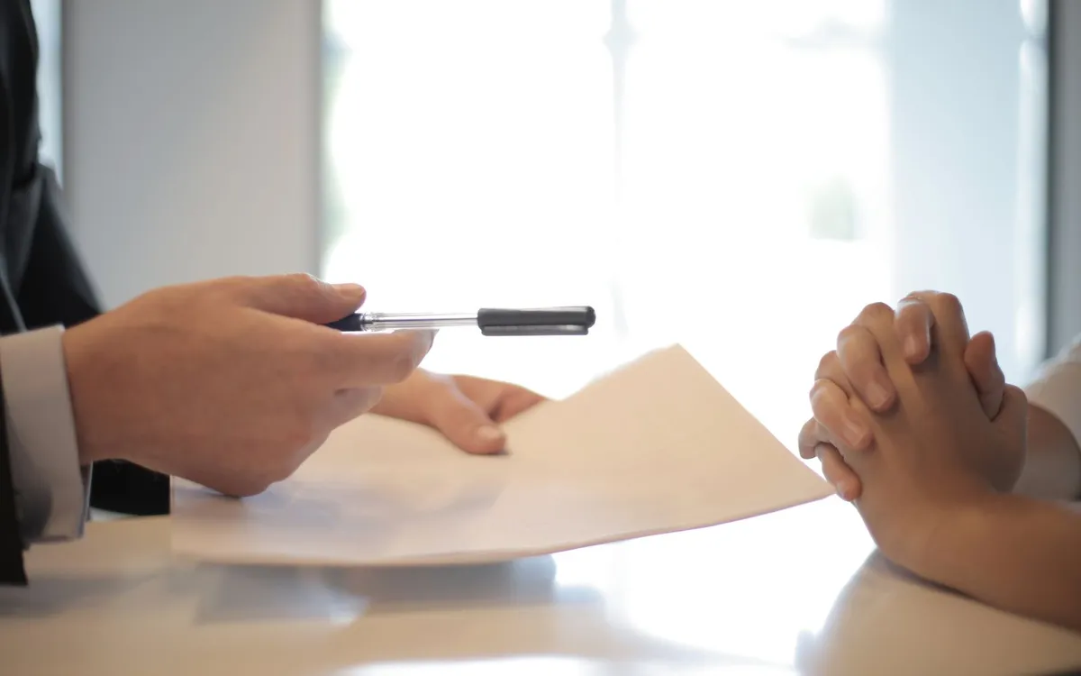 Mãos entregando um documento sobre a mesa durante assinatura de contrato.
