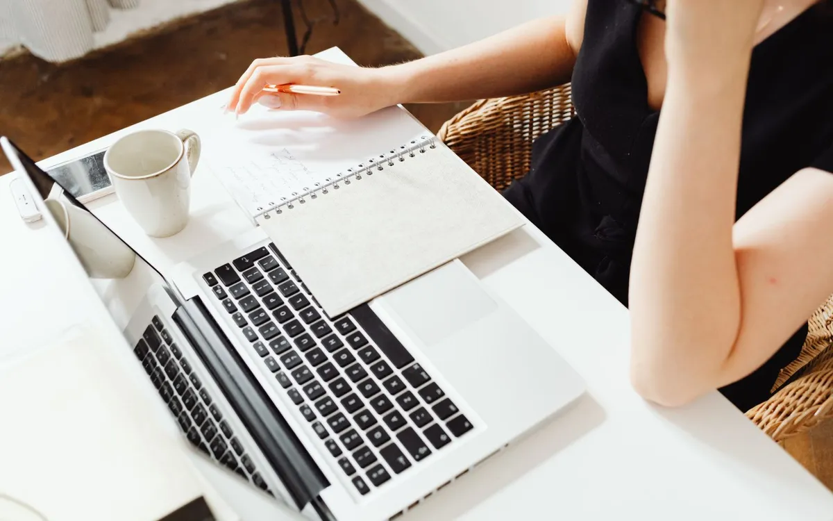 Mulher trabalhando no notebook com um caderno e uma xícara de café sobre a mesa.