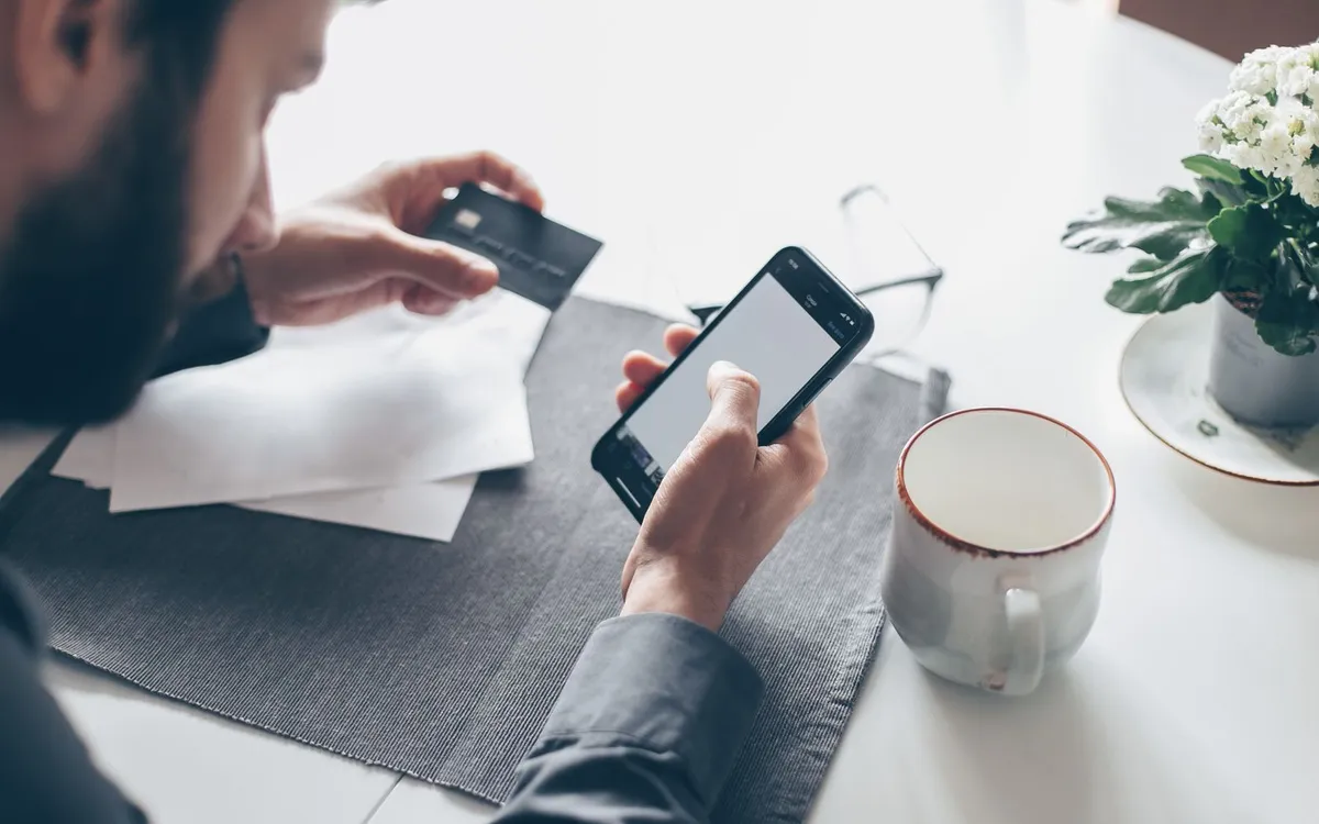 Homem conferindo o celular e um cartão sobre uma mesa com café e papéis.