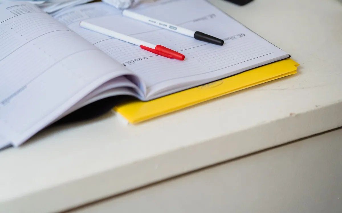 Caderno aberto sobre a mesa com canetas e um bloco amarelo.