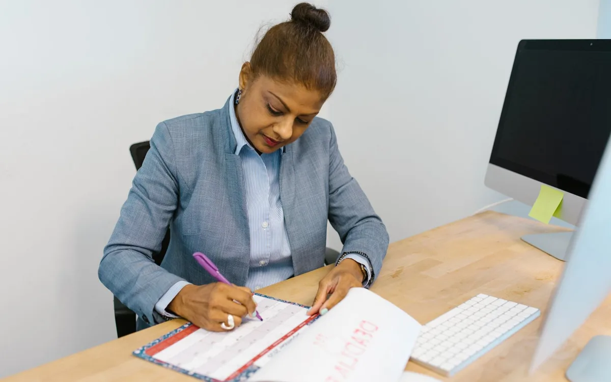 Mulher à mesa escrevendo em uma agenda, ao lado do computador, em um escritório.