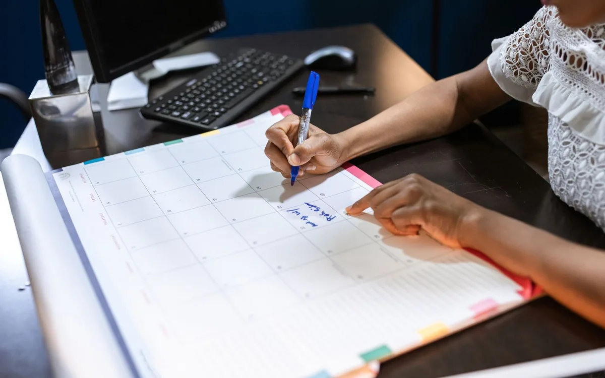 Mãos escrevendo em uma agenda grande sobre a mesa, ao lado do computador.