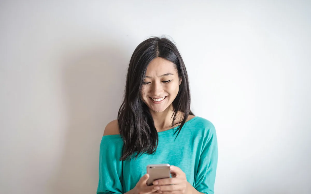 Mulher sorrindo ao olhar o celular, encostada em uma parede branca.