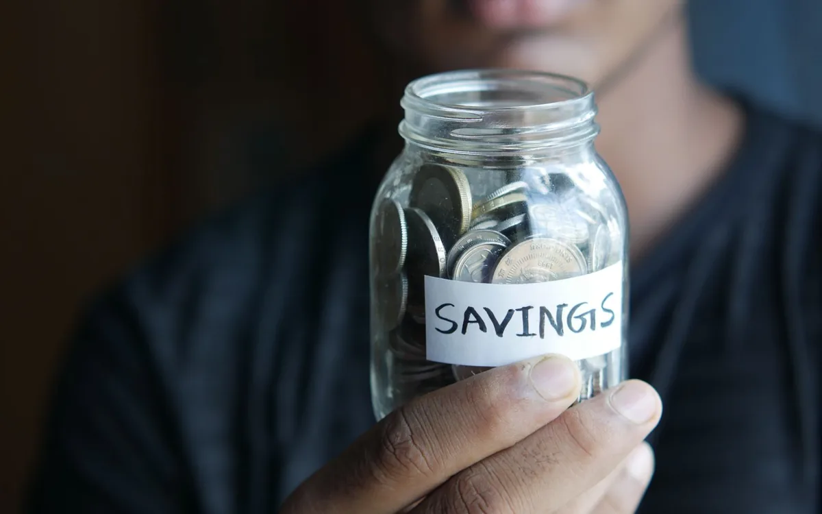 Pote de vidro com moedas e a palavra "savings" (economias), segurado por uma pessoa.