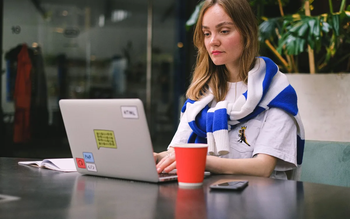 Jovem trabalhando em um notebook em uma mesa de café, com copo vermelho ao lado.