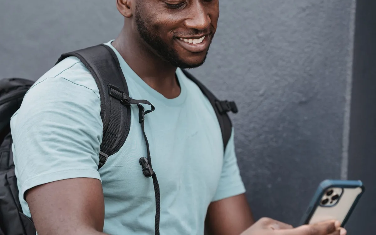 Homem sorrindo olhando o celular, com mochila nas costas, em uma calçada.