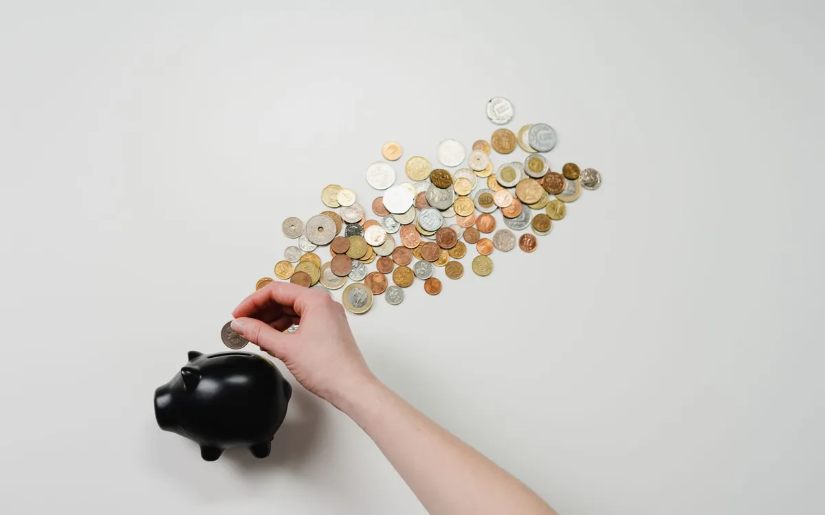 Mão colocando moedas em um cofre de porquinho preto sobre fundo claro.