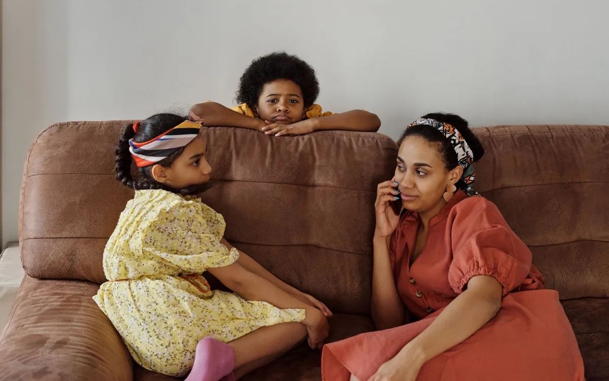 Mãe e filhos sentados em um sofá marrom, com a mulher ao telefone e as crianças ao lado.