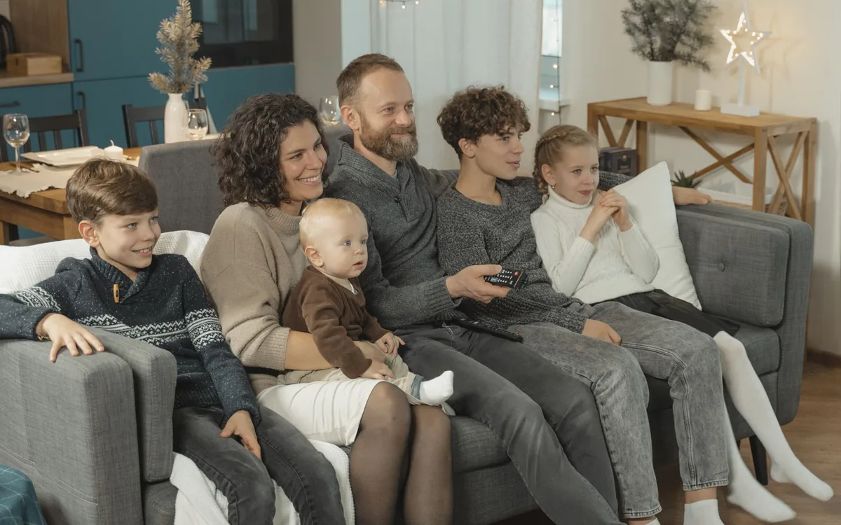 Família sentada em um sofá assistindo à televisão, com pai, mãe e filhos juntos.