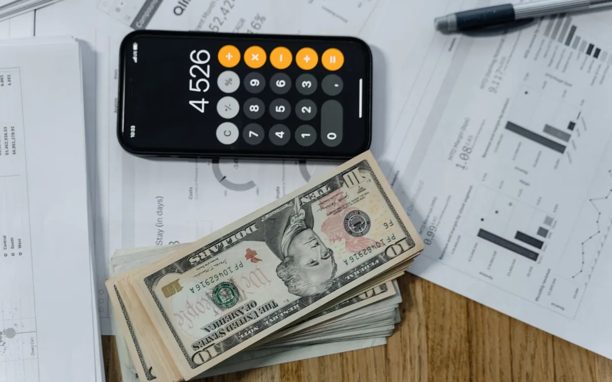 Celular com calculadora sobre notas de dinheiro e planilhas impressas.